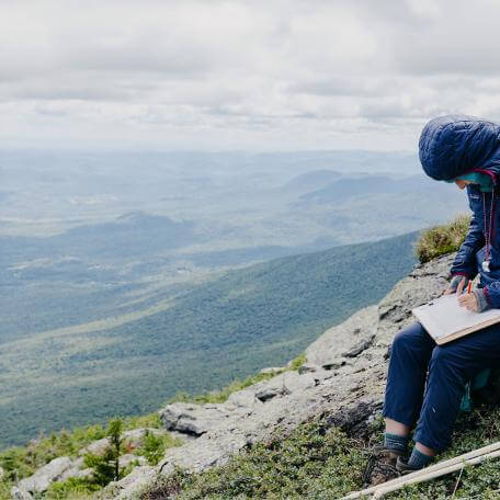 a student in a puffy jacket studies on a mountain side with a sweeping vista in the background