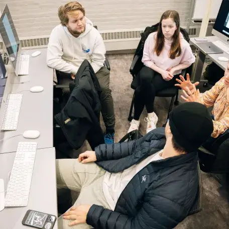 A group of students all sitting in chairs around a computer, listen to one student who is explaining something.