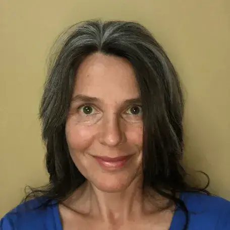 Liz, smiling for the camera, in an indoor setting, against a mustard colored background