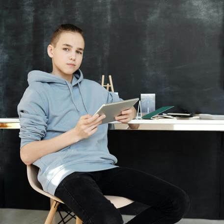 young teenager sitting at a desk behind a blackboard holding a tablet
