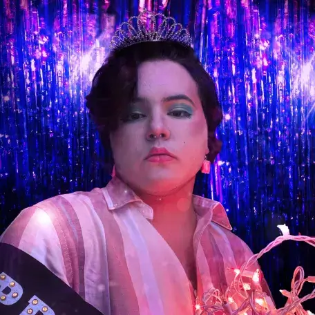 Juneau stands wearing a prom sash and holding string lights in front of a purple background. They wear a crown and green eyshadow in a pink and white striped shirt.