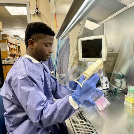 Current Student, Isreal, using the TC hood. Short haired male holding pipette wearing blue gown.