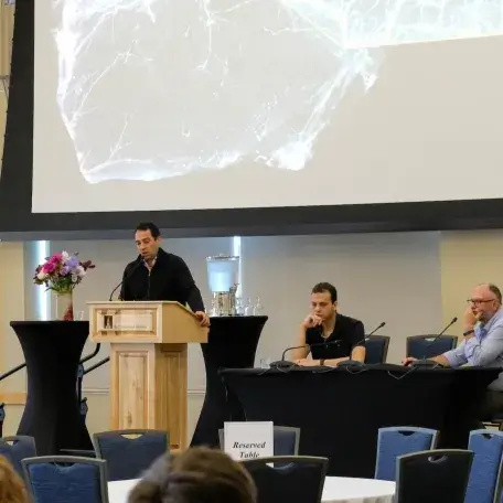 A researcher delivers a presentation at a podium in front of a large display screen, while two additional researchers are seated at a table to the side linking to Events Page