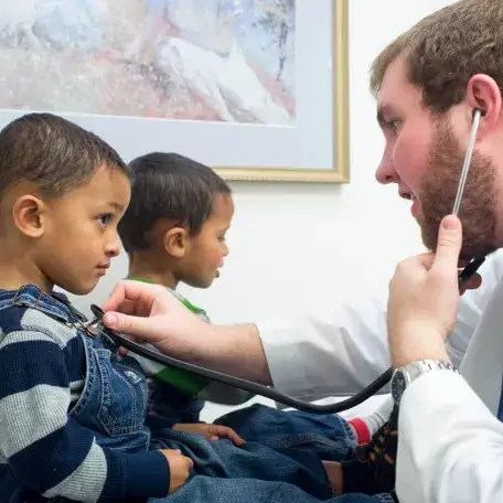 two children in a doctors office with a doctor using stethoscope to listen to ones heart