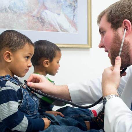 two children in a doctors office with a doctor using stethoscope to listen to ones heart