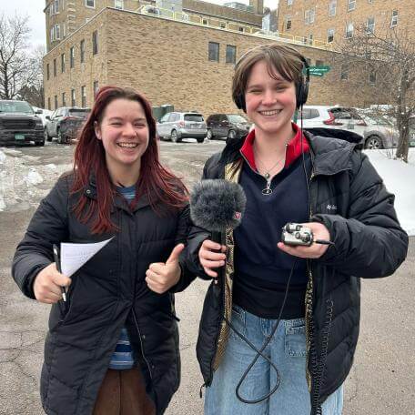 Two university of Vermont students wearing coats hold a microphone and notes while they cover the 2024 federal election.