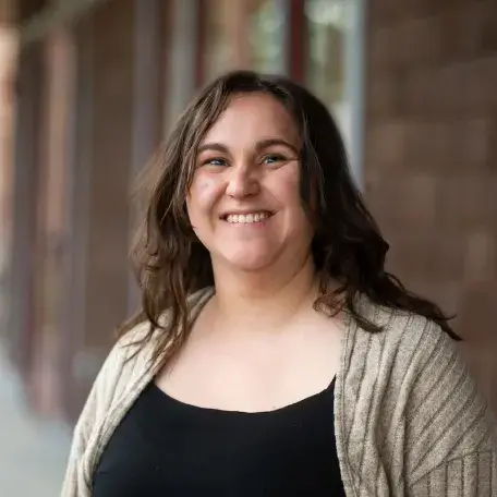 Kaitlyn, a woman with long wavy brown hair wearing a black top and cardigan, smiling in an outdoor headshot.
