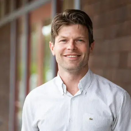 Tom, a man with short brown hair wearing a light button-down shirt, smiling in an outdoor headshot.