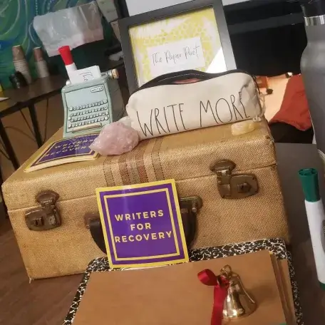 A tabletop display featuring a vintage typewriter, a decorative case labeled "WRITE MORE," a crystal, a sign that reads "WRITERS FOR RECOVERY," a brown notebook, a small bell, and a water bottle.