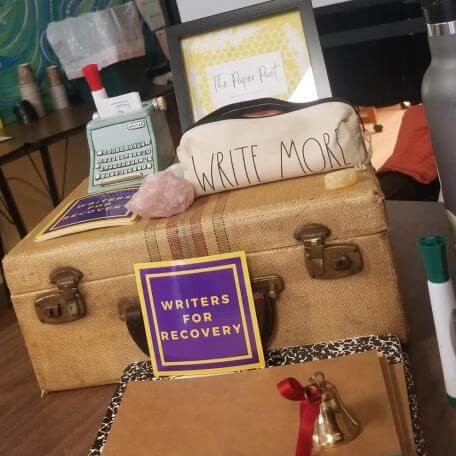 A tabletop display featuring a vintage typewriter, a decorative case labeled "WRITE MORE," a crystal, a sign that reads "WRITERS FOR RECOVERY," a brown notebook, a small bell, and a water bottle.
