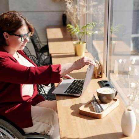 A white woman in her twenties with glasses and curly chestnut hair sits in a wheelchair, setting up a laptop at a café.