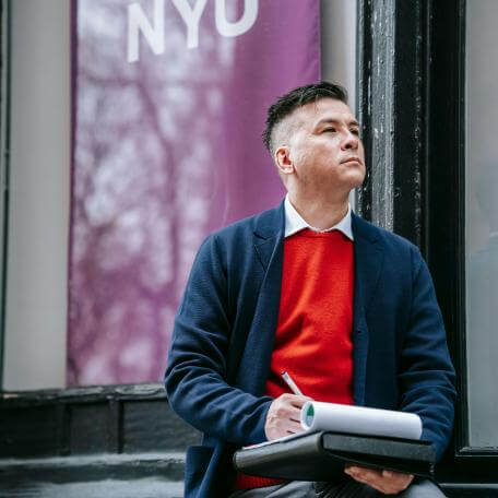 A Latino man in a red sweater and sport jacket stops to ponder while taking notes on a pad in his lap