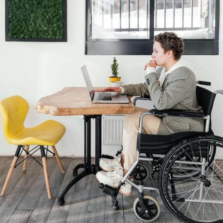A white college-aged person sits in a wheelchair at a table in front of an open laptop