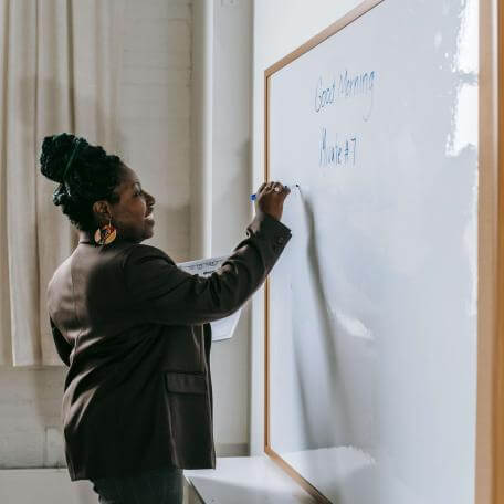 A Black female professor in a suit jacket, with braids piled into a bun atop her head, writes on a whiteboard in a classroom