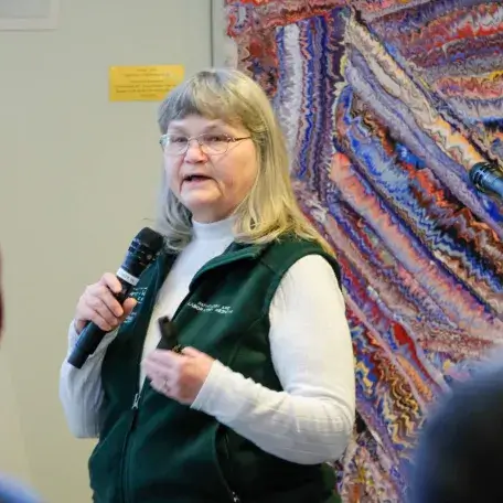 Individual wearing a green vest, white sweater, holding a microphone giving a presentation