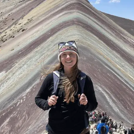 Person smiling in front of Mountain, with colorful layered slopes and hikers in the background.