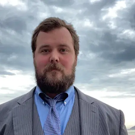 Person wearing a suit and tie standing outdoors near a rocky coastline under a cloudy sky.