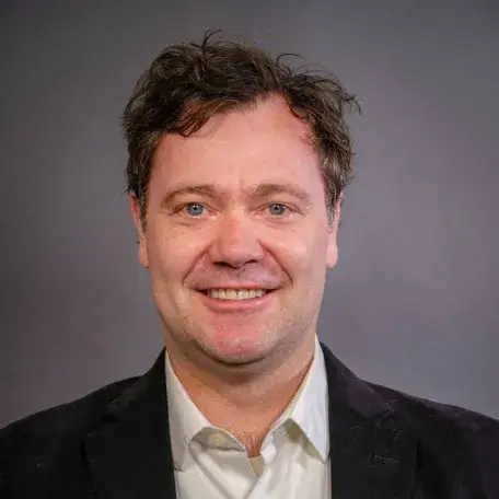 A photo of an individual standing in front of a gray background, who has brown hair and blue eyes, wearing a button down light gray shirt with a black sport coat.