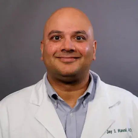A photo of a smiling, bald-headed individual in a light blue button-down shirt and white doctor's coat, standing in front of a gray background.