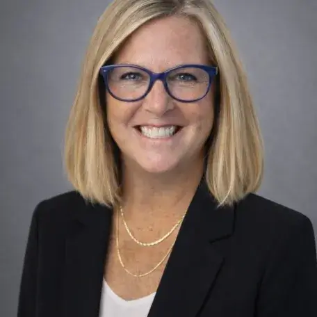 Sharon Mone: pale-skinned woman with a blonde sidepart bob and glasses, in a black blazer and white shell
