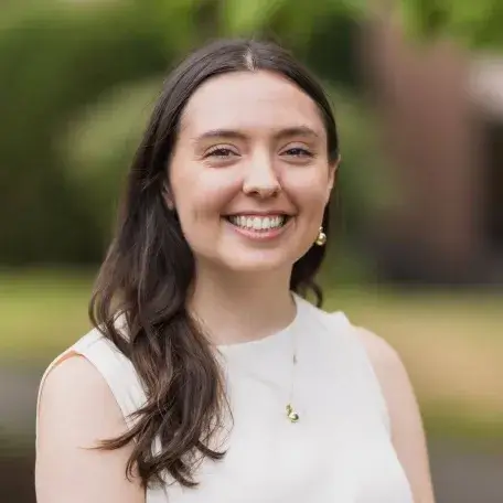 Green tree background, Julia is smiling looking into camera. She is wearing beige top