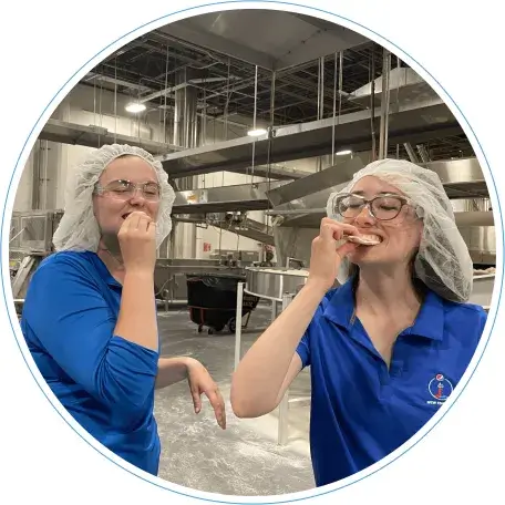 Two student interns taste a cookie while standing at a lays factory floor, wearing hair and eye protection