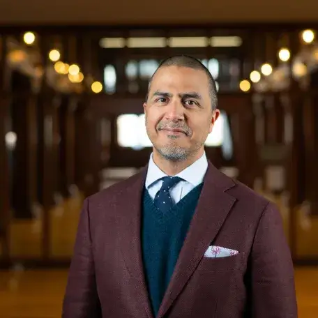 ahmed in a burgundy suit with a navy tie, looking straight ahead and smiling in front of a blurred background.