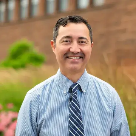 Jorge is pictured wearing a blue striped shirt and a blue patterned tie posed outdoors, with pink flowers and a brick building softly blurred behind him. 