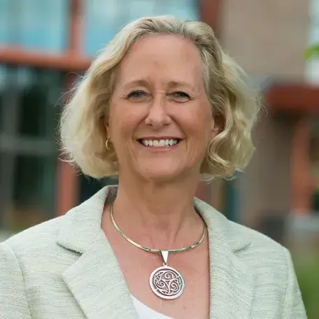 Nancy Mathews standing outside the Aiken Center at UVM, she is wearing a large silver necklace, a beige blazer, and has chin-length blonde hair. She is smiling.