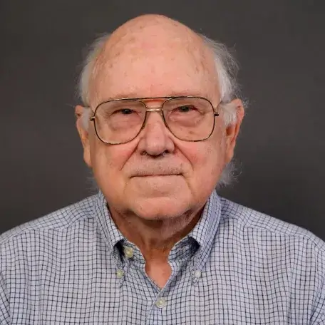 Male balding with salt and pepper hair around the sides of his head, wearing glasses and a white and blue gingham patterned shirt.