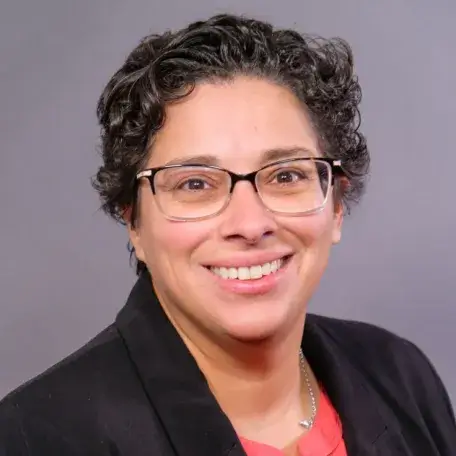 Smiling woman with short curly black hair wearing glasses and a black blazer over a red blouse.