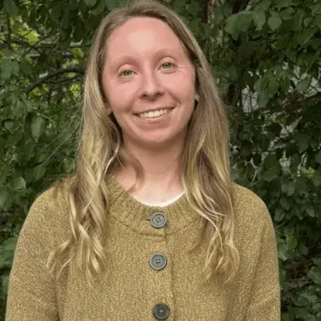 Nicole posing in front of background of green leaves. Nicole is in the center, smiling, and wearing a dark beige cardigan with chunky black buttons. 