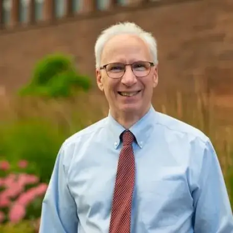 A man with white hair and glasses wearing a blue button down shirt with a red tie photographed outside against a red stone building.