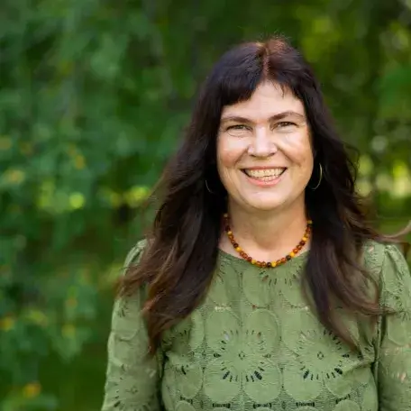 A woman with long dark hair is standing in front of trees