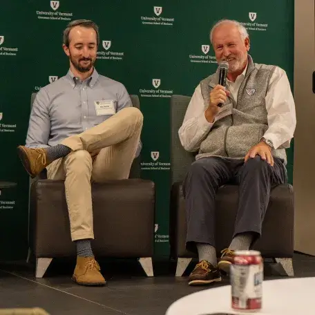 L to R: Mac Beitzel and Skip Beitzel (’76) sit on stage while Skip speaks into a mic