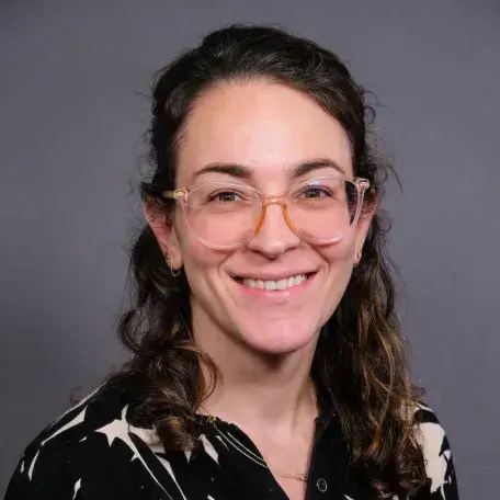 A person with long dark hair and glasses wearing a black patterned blouse