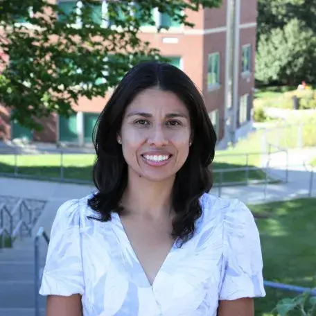 Laura Sanchez-Parkinson smiling in a white shirt with green foliage and a brick building behind her.  