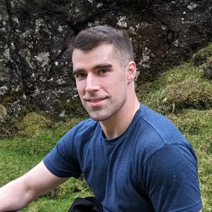 A pale-skinned man in his early thirties, with close-cropped dark brown hair, dark brown eyes, and light stubble.