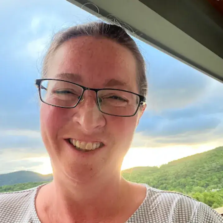 Middle-aged white woman wearing glasses stands in front of sunrise over Vermont mountains.