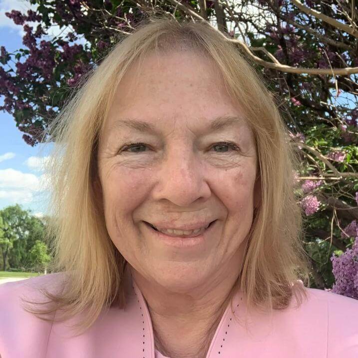 A light skinned woman with a pink cardigan stands in front of a tree, she is smiling.