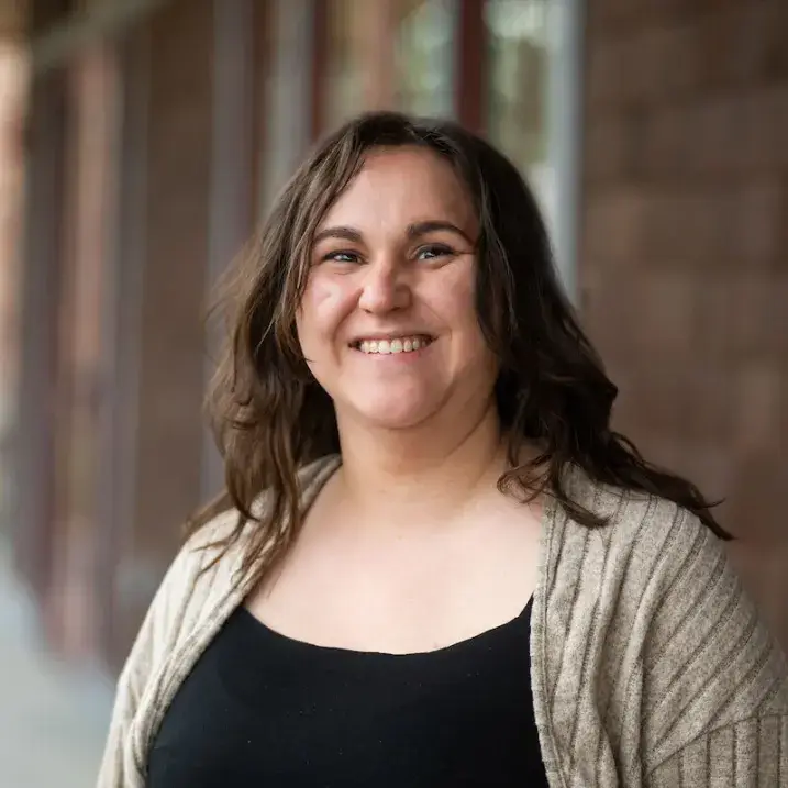 Kaitlyn, a woman with long wavy brown hair wearing a black top and cardigan, smiling in an outdoor headshot.