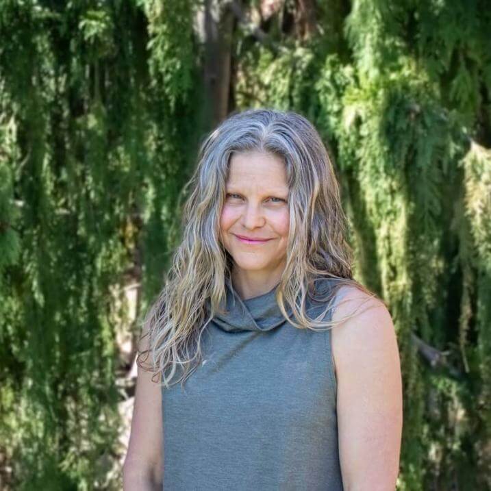 Rachelle Gould headshot, she is wearing a grey sleeveless shirt and is standing in front of a green, forested background