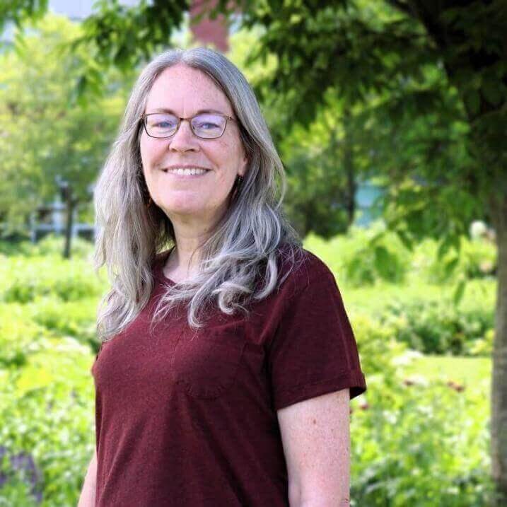 A woman with glasses and a smiling in from of a verdant background.