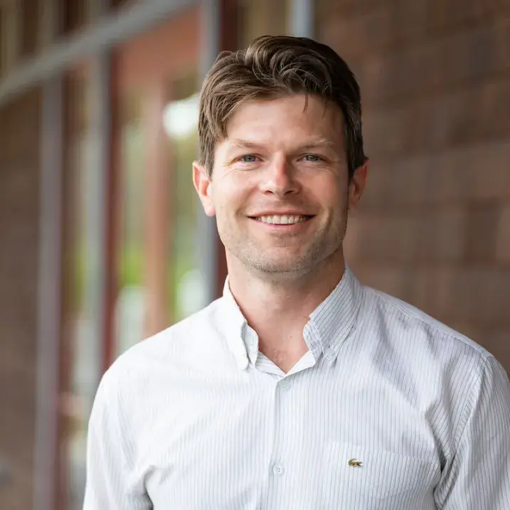 Tom, a man with short brown hair wearing a light button-down shirt, smiling in an outdoor headshot.