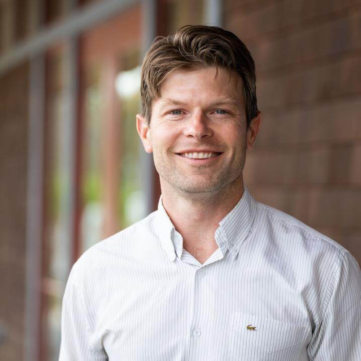 Tom, a man with short brown hair wearing a light button-down shirt, smiling in an outdoor headshot.