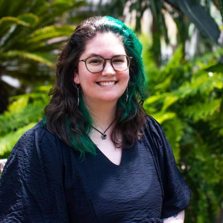 Headshot of Hannah Kershaw, smiling, sitting in front of greenery, wearing a black shirt and glasses.