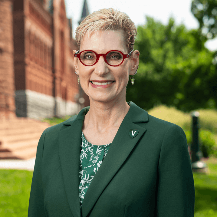 a person wearing red glasses and a green blazer poses in front of red brick buildings