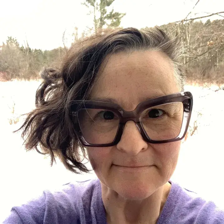 White non-binary person in their forties with wild short dark hair and large, heavy-framed glasses.