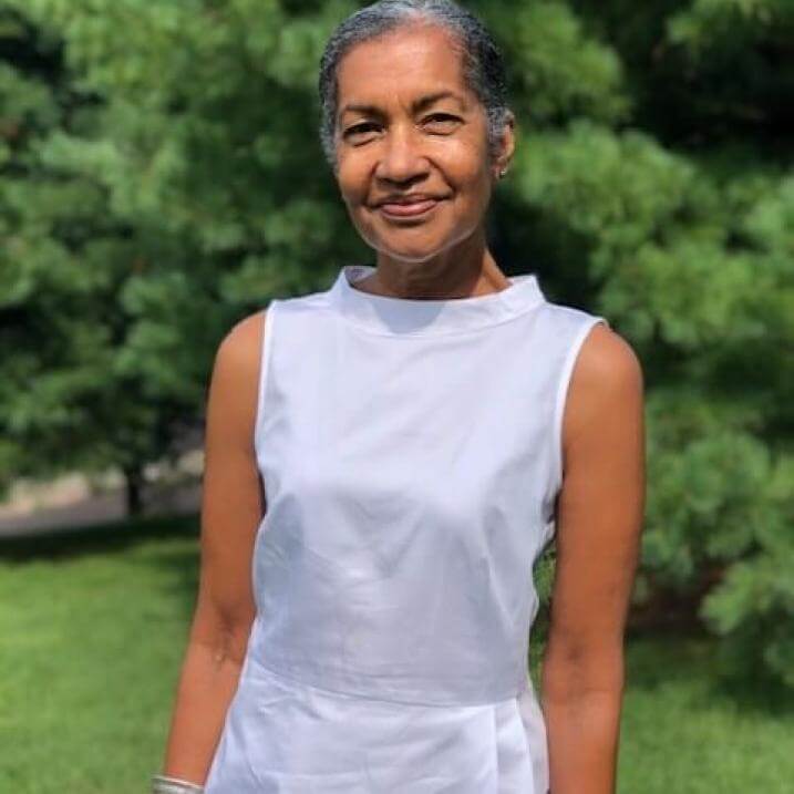 Photo of Bev smiling , wearing a white blouse with white pine trees in the background