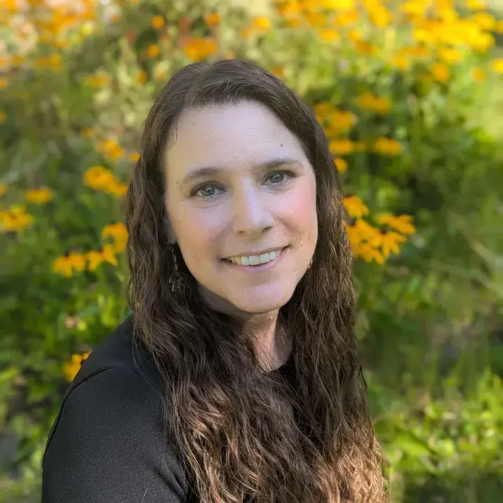 Julie Chiappinelli headshot: pale-skinned woman with long dark wavy hair and dark eyes, looking straight into the camera in a field of tall yellow flowers.